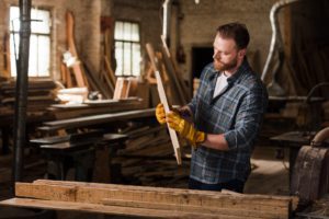 bearded carpenter in protective gloves working with wood at sawmill