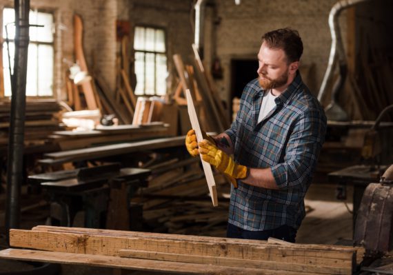 bearded carpenter in protective gloves working with wood at sawmill