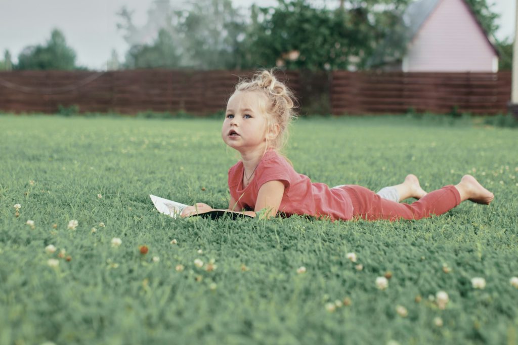 little blonde girl reading a book and lying on the lawn